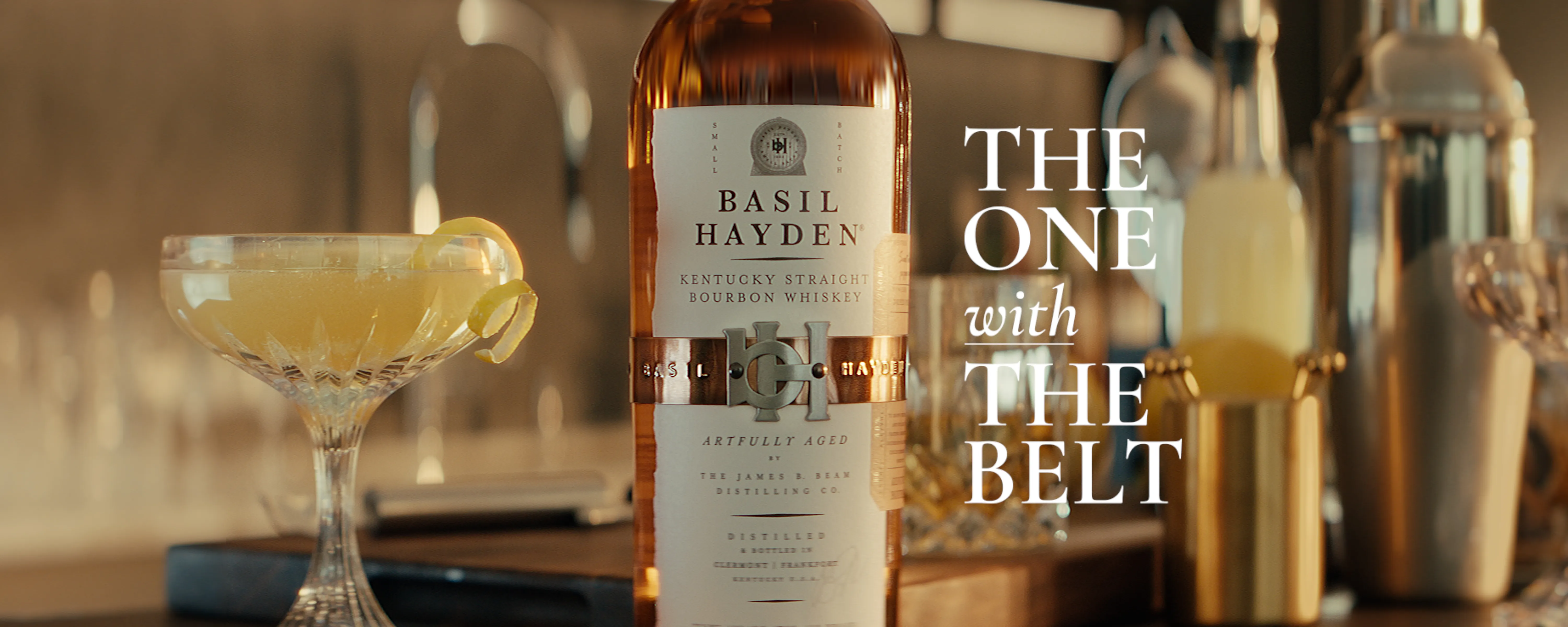 A bottle of Basil Hayden bourbon and a cocktail with a lemon twist on a wooden bar, with text overlay promoting the brand.